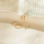 Set of gold hoop earrings on a soft beige surface with a glass container in the background.